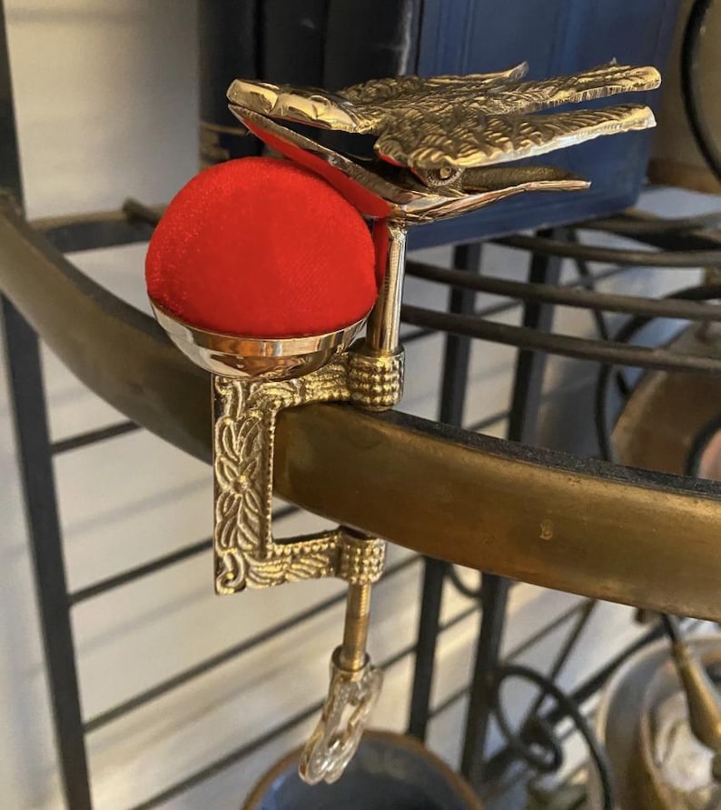 May include: A brass sewing clamp featuring an embossed floral pattern on the vertical arm and a detailed bird figure at the top. A bright red, spherical fabric pincushion is mounted in a polished brass cup attached to the side of the metal clamp.