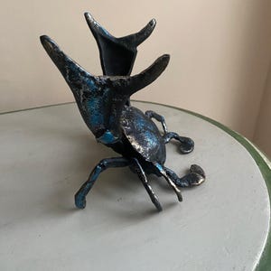 May include: A detailed sculpture of a beetle with a metallic finish. The beetle is primarily dark with blue and silver accents, and features prominent antennae and legs. The sculpture is displayed on a light gray surface.