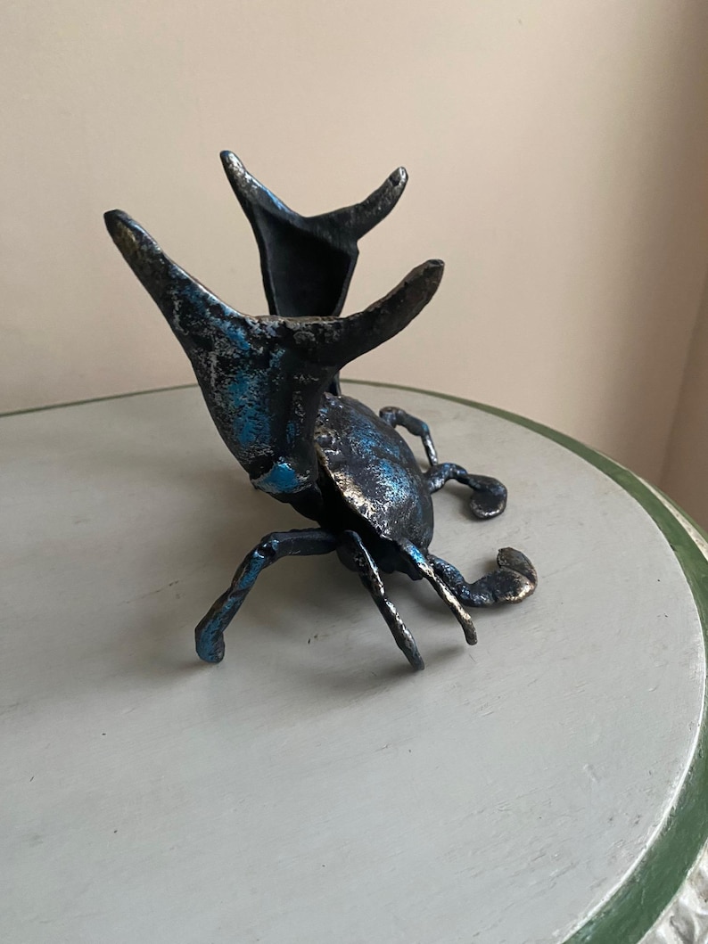 May include: A detailed sculpture of a beetle with a metallic finish. The beetle is primarily dark with blue and silver accents, and features prominent antennae and legs. The sculpture is displayed on a light gray surface.