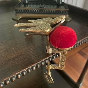 May include: A brass sewing clamp featuring a bird-shaped top with detailed wing feathers and a bright red velvet pincushion. The metal clamp mechanism includes an adjustable screw with a decorative handle, designed to attach to a table edge.