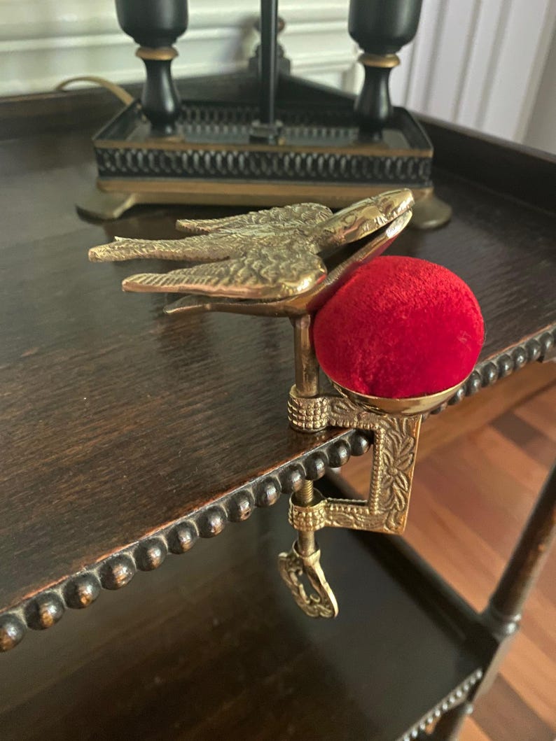 May include: A brass sewing clamp featuring a bird-shaped top with detailed wing feathers and a bright red velvet pincushion. The metal clamp mechanism includes an adjustable screw with a decorative handle, designed to attach to a table edge.