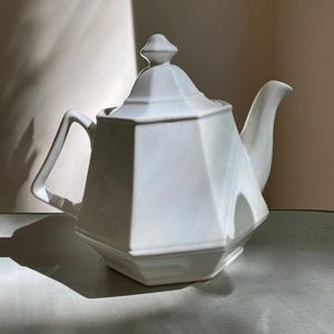 Könnte beinhalten: Eine weiße Porzellan-Teekanne mit geometrischem Design. Die Teekanne hat einen Deckel mit kleinem Knauf, einen gebogenen Ausguss und einen eckigen Griff. Die Teekanne steht auf einer hellen Oberfläche.