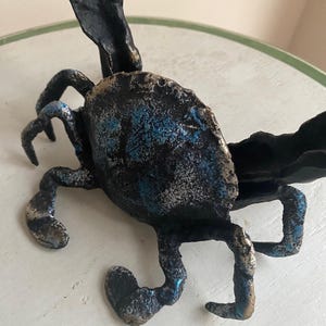May include: A handcrafted metal sculpture of a crab with a textured surface. The crab is primarily black with accents of blue and silver. The sculpture has detailed legs and claws, and a large, textured body.