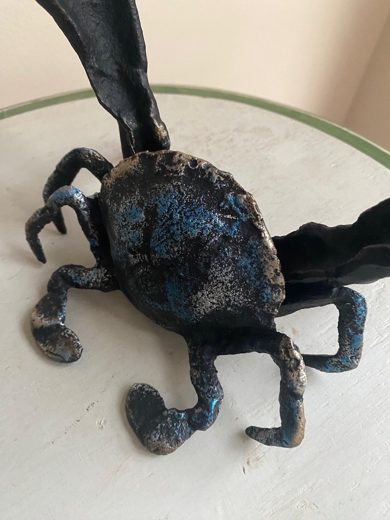 May include: A handcrafted metal sculpture of a crab with a textured surface. The crab is primarily black with accents of blue and silver. The sculpture has detailed legs and claws, and a large, textured body.
