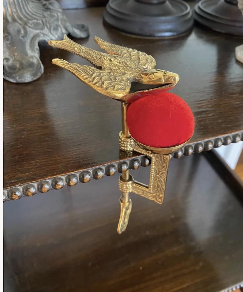 May include: A brass sewing clamp featuring a bird figure with detailed wing engravings and a bright red circular velvet pincushion. The metal clamp mechanism includes an adjustable screw with a decorative handle and an embossed floral pattern.