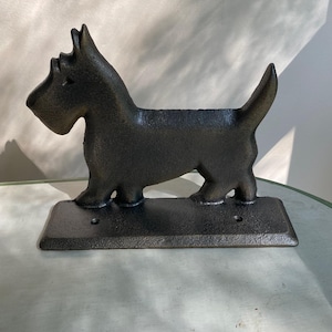 May include: A dark gray cast iron Scottie dog figurine. The dog is standing on a rectangular base with two holes. The figurine has a textured surface and is a decorative item.