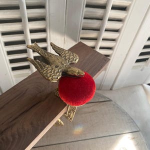 May include: A brass bird-shaped sewing clamp featuring a textured wing design and a bright red velvet pincushion. The metal clamp is attached to a wooden board, showcasing an ornate floral pattern engraved along the side of the metal hardware.