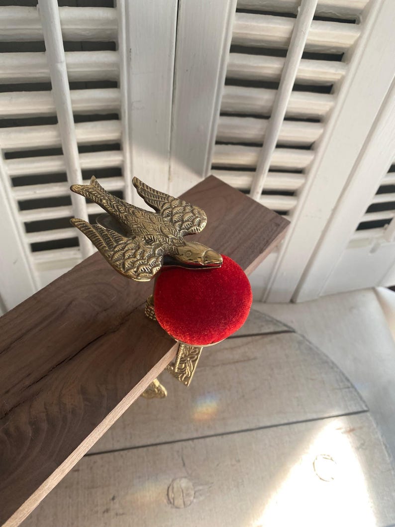 May include: A brass bird-shaped sewing clamp featuring a textured wing design and a bright red velvet pincushion. The metal clamp is attached to a wooden board, showcasing an ornate floral pattern engraved along the side of the metal hardware.