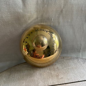 May include: A shiny, gold-colored bell with a rounded top and a small handle. The bell has the inscription "A bell is not a bell till you ring it." in black lettering. The bell is resting on a light-colored wooden surface.