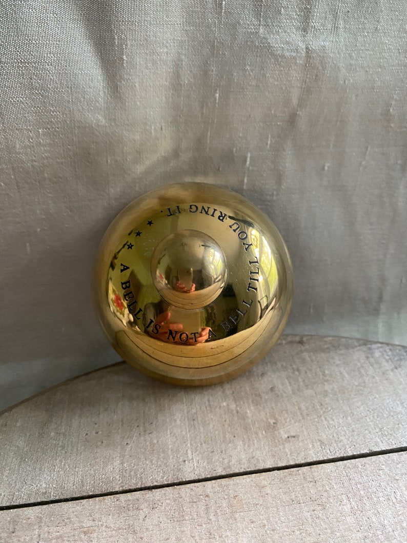 May include: A shiny, gold-colored bell with a rounded top and a small handle. The bell has the inscription "A bell is not a bell till you ring it." in black lettering. The bell is resting on a light-colored wooden surface.