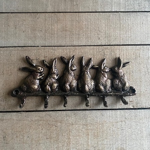 May include: Bronze-colored metal wall hook featuring six rabbit figures. Each rabbit is perched on a horizontal bar, with a hook extending below each one. The decorative piece is mounted on a wooden surface.