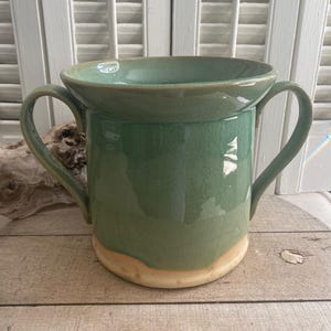 May include: A sage green ceramic vessel featuring a wide, flared rim and two curved handles on opposite sides. The glaze has a subtle crackle texture, and the base of the pot is exposed, showing a light beige, unglazed clay finish.