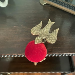 May include: A brass bird-shaped clamp with detailed wing engravings attached to a round, bright red velvet pincushion. The metal bird features a textured surface and is positioned with its wings spread, securely holding the circular red cushion.