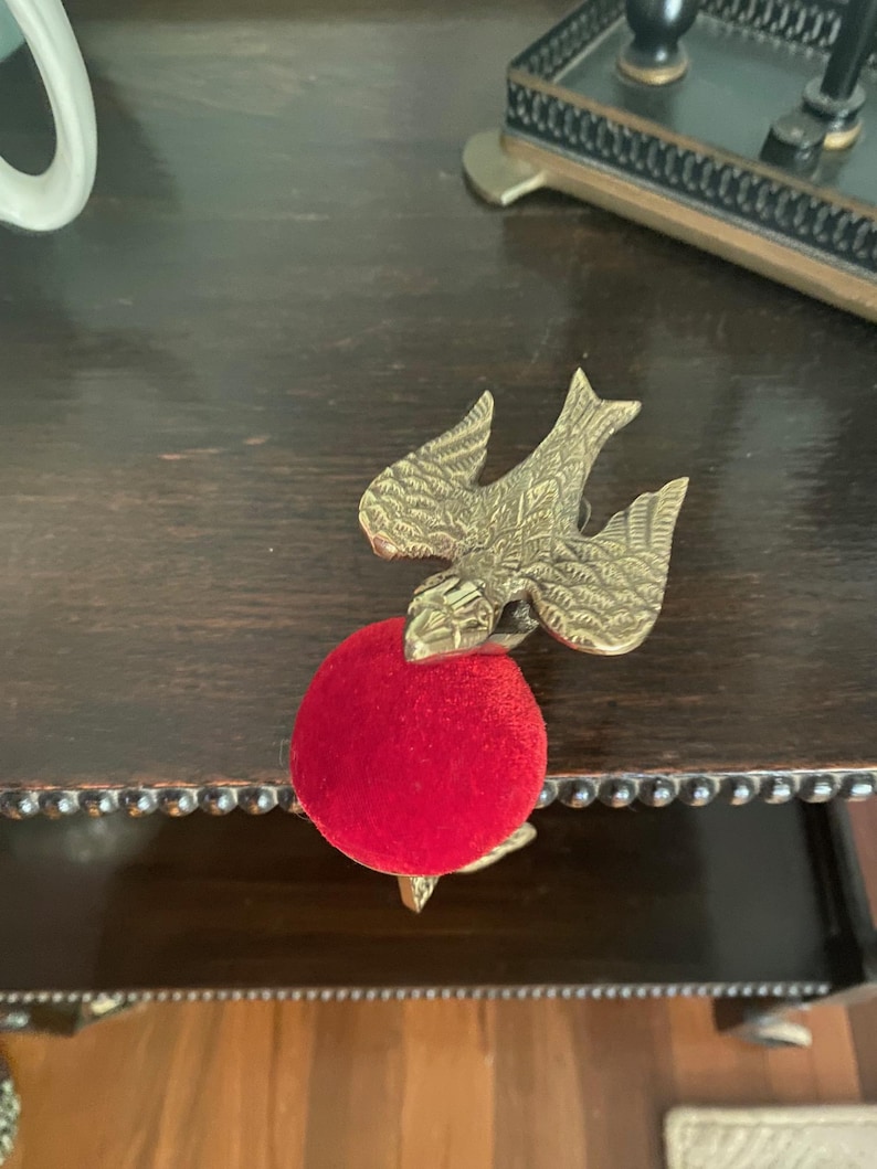 May include: A brass bird-shaped clamp with detailed wing engravings attached to a round, bright red velvet pincushion. The metal bird features a textured surface and is positioned with its wings spread, securely holding the circular red cushion.