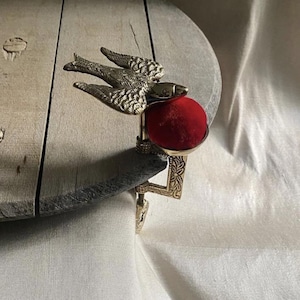 May include: A brass clamp-on pincushion featuring a detailed bird figure with outstretched wings. The device includes a vibrant red velvet cushion held within a metal cup, attached to a decorative metal screw clamp for securing to a table edge.