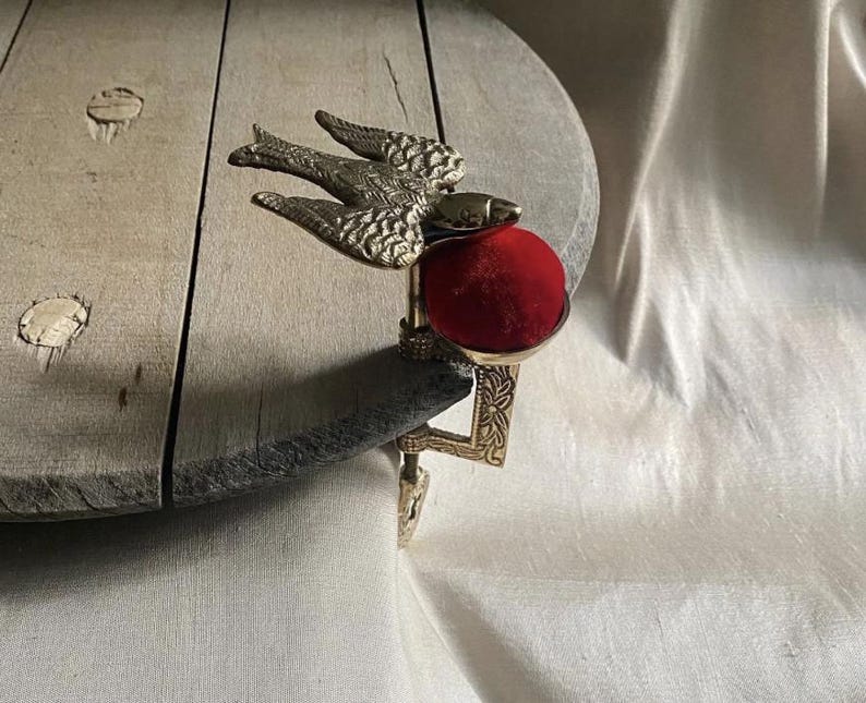 May include: A brass clamp-on pincushion featuring a detailed bird figure with outstretched wings. The device includes a vibrant red velvet cushion held within a metal cup, attached to a decorative metal screw clamp for securing to a table edge.