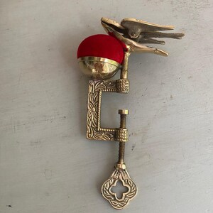 May include: A brass sewing clamp featuring an ornate C-shaped frame with floral engravings and a threaded tightening screw. A bright red velvet pincushion sits atop the clamp, accompanied by a decorative brass bird figure perched on the side.