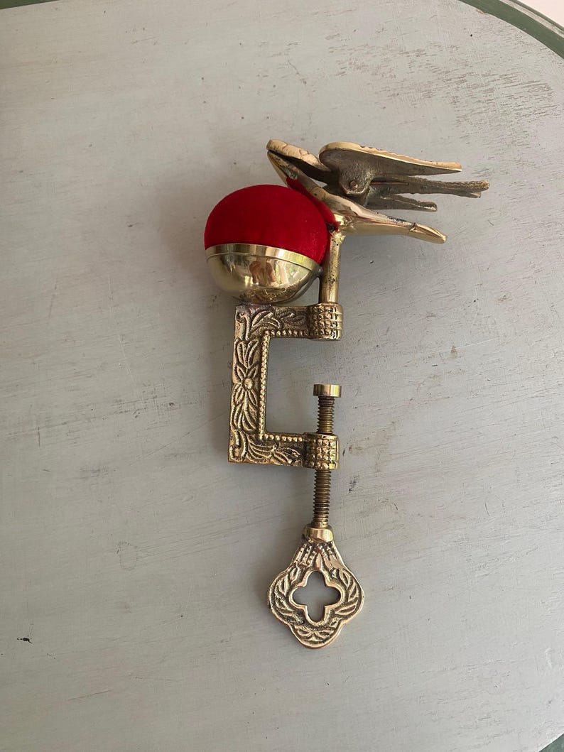 May include: A brass sewing clamp featuring an ornate C-shaped frame with floral engravings and a threaded tightening screw. A bright red velvet pincushion sits atop the clamp, accompanied by a decorative brass bird figure perched on the side.
