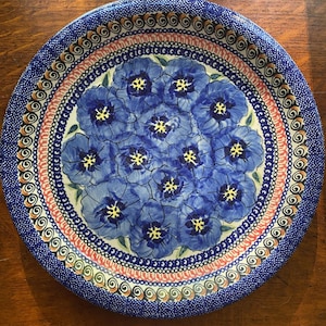 May include: A round, ceramic plate with a blue floral design. The plate features a central cluster of blue flowers with yellow centers, surrounded by decorative borders in blue, pink, and brown. The plate is suitable for serving food.