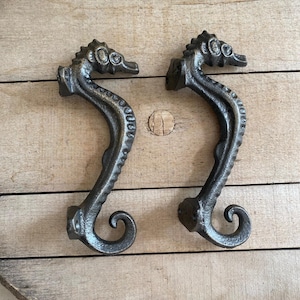 May include: Two cast iron seahorse-shaped cabinet pulls. The pulls are dark gray with detailed textures, including the seahorse's head, body, and tail. The pulls are mounted on a light brown wooden surface.
