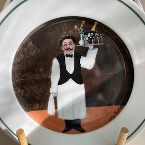 May include: White ceramic plate with a green border, featuring a waiter illustration carrying champagne and flowers. The waiter wears a black waistcoat and white apron. The plate is displayed on a wooden stand, suitable for decorative use.