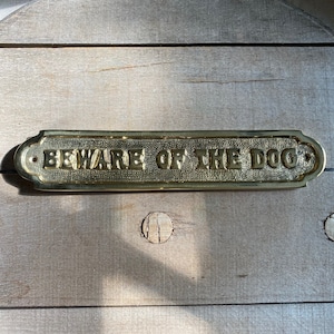 以下が含まれることがあります： 丸みを帯びた縁と斜めの表面を持つ金色の金属製の標識。標識には「BEWARE OF THE DOG」という文字が浮き彫りにされています。