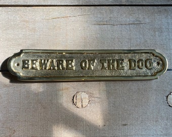 May include: A gold-colored metal sign with rounded edges and a beveled surface. The sign reads "BEWARE OF THE DOG" in raised letters.
