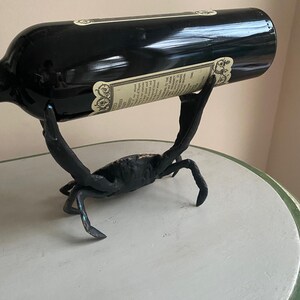 May include: A black wine bottle rests in a crab-shaped metal wine holder. The crab is black with detailed legs and claws. The bottle has a label with text. The holder is on a light green and white table.