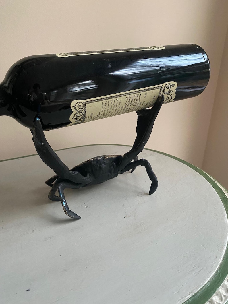 May include: A black wine bottle rests in a crab-shaped metal wine holder. The crab is black with detailed legs and claws. The bottle has a label with text. The holder is on a light green and white table.