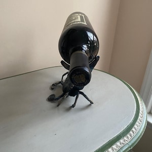 May include: A dark wine bottle is held by a decorative metal scorpion wine holder. The scorpion is dark gray with detailed legs and claws. The bottle is resting on a white and green painted table.