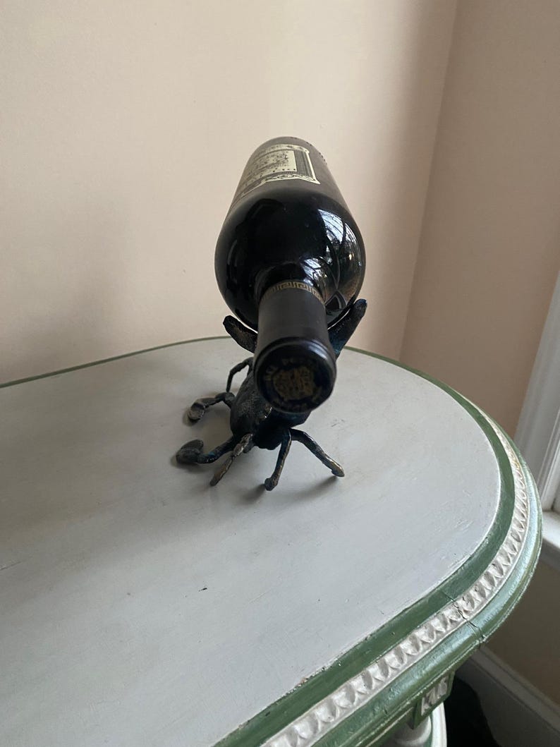 May include: A dark wine bottle is held by a decorative metal scorpion wine holder. The scorpion is dark gray with detailed legs and claws. The bottle is resting on a white and green painted table.