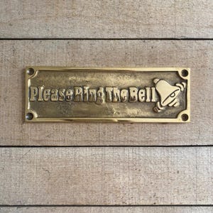 May include: A rectangular brass door sign with the words "Please Ring The Bell" in embossed lettering. A bell graphic is on the right side. The sign has four screw holes and is set against a wooden background.