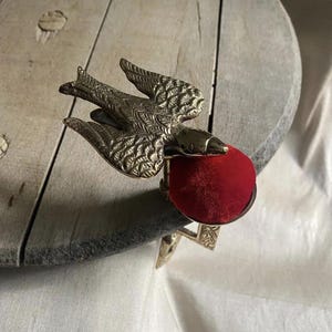 May include: A brass bird-shaped sewing clamp featuring a textured wing design and a circular metal base topped with a vibrant red velvet pincushion. The metal bird is positioned with its wings spread, attached to a sturdy clamp mechanism.