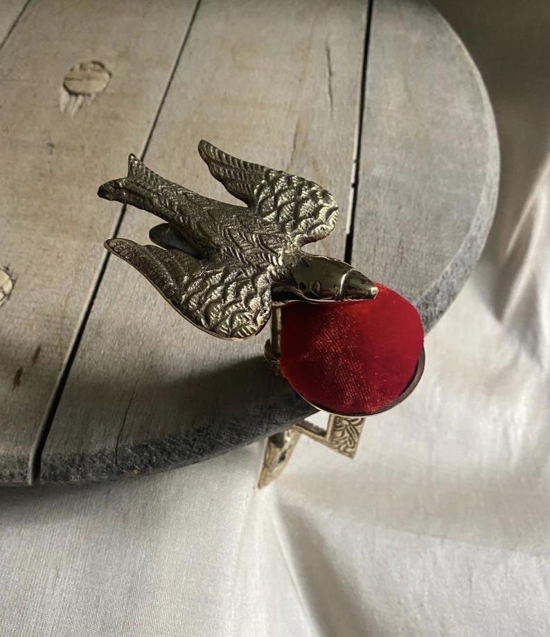 May include: A brass bird-shaped sewing clamp featuring a textured wing design and a circular metal base topped with a vibrant red velvet pincushion. The metal bird is positioned with its wings spread, attached to a sturdy clamp mechanism.
