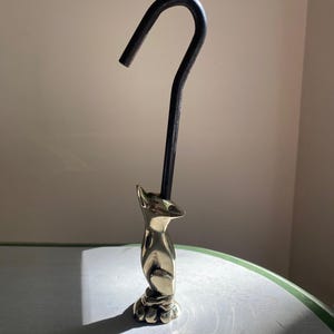 May include: A metallic, gold-toned fox figurine standing on a base, featuring a long, slender, black curved hook extending upward from the top of the fox's head. The fox is depicted in a seated position with its head tilted slightly upward.