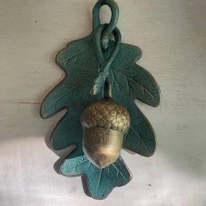 May include: A decorative wall hanging featuring a textured, verdigris-colored oak leaf with a prominent, metallic gold-toned acorn suspended from the top loop. The piece has a weathered, aged appearance with visible veins on the leaf and a detailed cap.