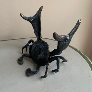 May include: A dark metal sculpture of a beetle with outstretched appendages. The beetle has a textured surface with hints of blue and bronze. The sculpture is placed on a white and green table.