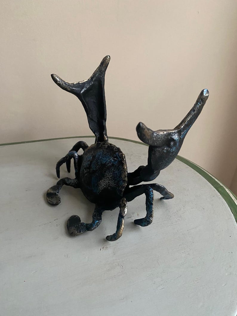 May include: A dark metal sculpture of a beetle with outstretched appendages. The beetle has a textured surface with hints of blue and bronze. The sculpture is placed on a white and green table.