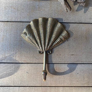 Peut inclure: Un crochet mural en forme de coquillage en laiton. Le crochet présente un design détaillé en forme d'éventail avec un long crochet incurvé s'étendant du centre. Le crochet est monté sur une surface en bois.