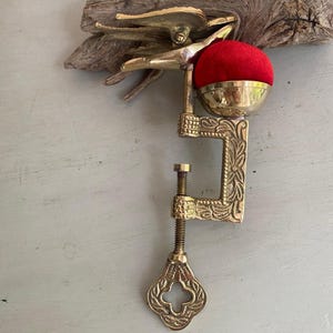 May include: A brass sewing clamp featuring an ornate C-shaped frame with floral engravings, a threaded tightening screw with a decorative handle, and a bird-shaped top. A bright red velvet pincushion is mounted in a brass cup on the upper arm.