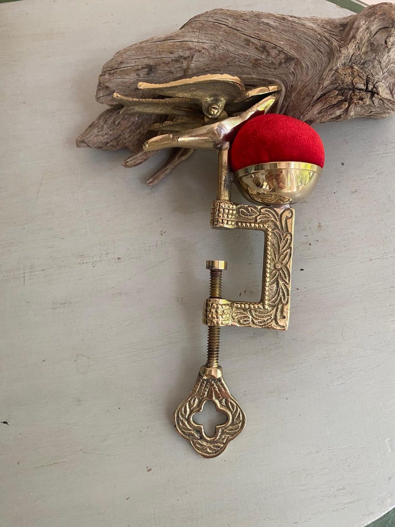 May include: A brass sewing clamp featuring an ornate C-shaped frame with floral engravings, a threaded tightening screw with a decorative handle, and a bird-shaped top. A bright red velvet pincushion is mounted in a brass cup on the upper arm.