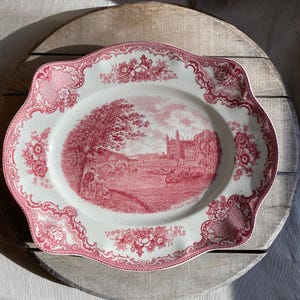 Puede incluir: Plato de servir ovalado y antiguo con un diseño floral y paisajístico rojo y blanco. El borde presenta un borde festoneado y detalles florales. El centro representa un paisaje escénico con figuras y edificios.