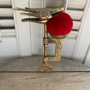 May include: A brass sewing clamp featuring a bird-shaped top clip and a bright red velvet pincushion. The metal clamp has an ornate floral pattern and a threaded screw mechanism for attachment, designed to hold fabric securely while sewing.