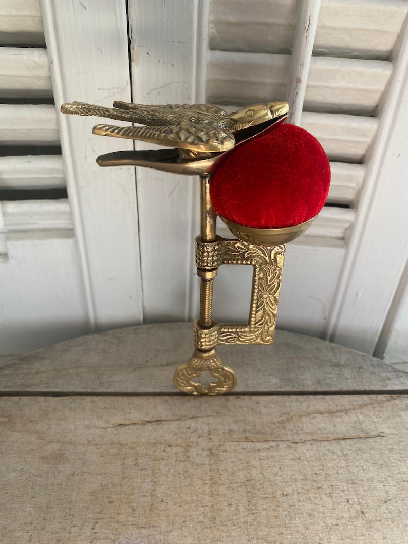 May include: A brass sewing clamp featuring a bird-shaped top clip and a bright red velvet pincushion. The metal clamp has an ornate floral pattern and a threaded screw mechanism for attachment, designed to hold fabric securely while sewing.