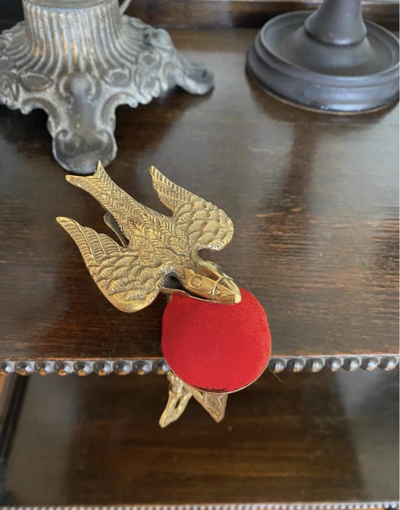 May include: A brass bird-shaped sewing clamp featuring detailed wing engravings and a circular red velvet pincushion attached to the front. The metal bird is positioned with its wings spread, serving as a functional tool for holding fabric.