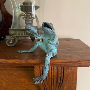 May include: A verdigris-colored metal frog figurine with a textured surface, depicted in a seated position with one leg extended forward. The frog has an open mouth and is holding a small, rectangular, book-like object in its front limbs.
