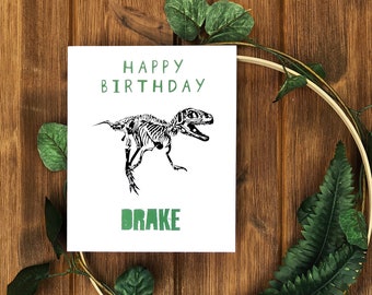 May include: A white card with green text that says "Happy Birthday" and "Drake". A black and white dinosaur skeleton illustration is in the center of the card.