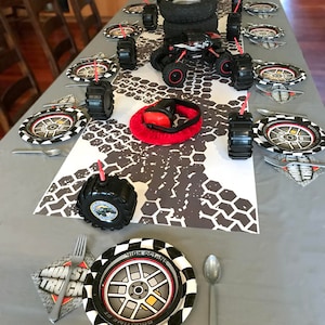 May include: A monster truck themed party table setting with black and white checkered plates, black tire-shaped cups, and a red monster truck toy. The table is covered in a gray tablecloth with a tire tread pattern.
