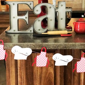 May include: A red stand mixer with a silver bowl and the word "KitchenAid" on the side. The mixer is on a countertop with a silver metal sign that says "Eat" and a red and white checkered apron hanging from a string.
