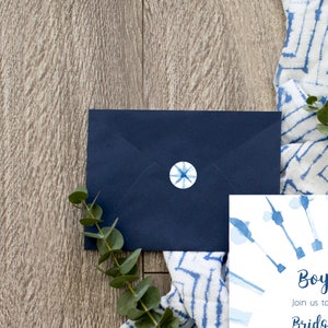 May include: A blue envelope with a white star sticker on the front. The envelope is lying on a wooden surface with a blue and white patterned fabric and a sprig of eucalyptus.